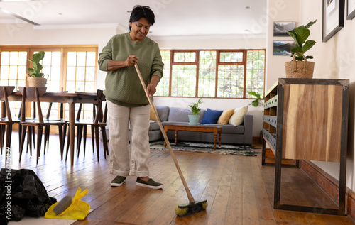 Full length of biracial mature woman with short hair sweeping hardwood floor with broom at home photo