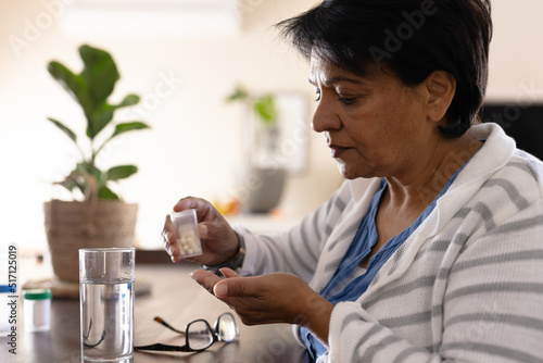 Biracial mature woman with drinking water and eyeglasses on table taking medicines at home photo