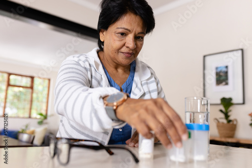 Biracial mature woman with medicine bottles and eyeglasses sitting on table at home, copy space photo