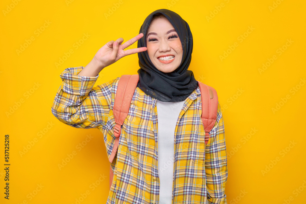 Smiling young Asian Muslim woman student in casual clothes and backpack, looking at camera, showing peace sign with fingers isolated on yellow background. Education school university college concept Smiling young Asian Muslim woman student in casual clothes and backpack, looking at camera, showing peace sign with fingers isolated on yellow background. Education school university college concept
