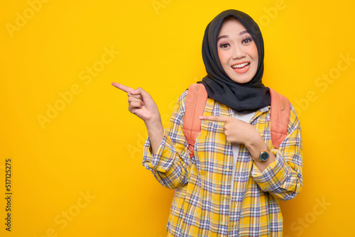 Cheerful young Asian Muslim woman student in casual clothes and backpack, pointing fingers at copy space isolated on yellow background.Education school university college concept
