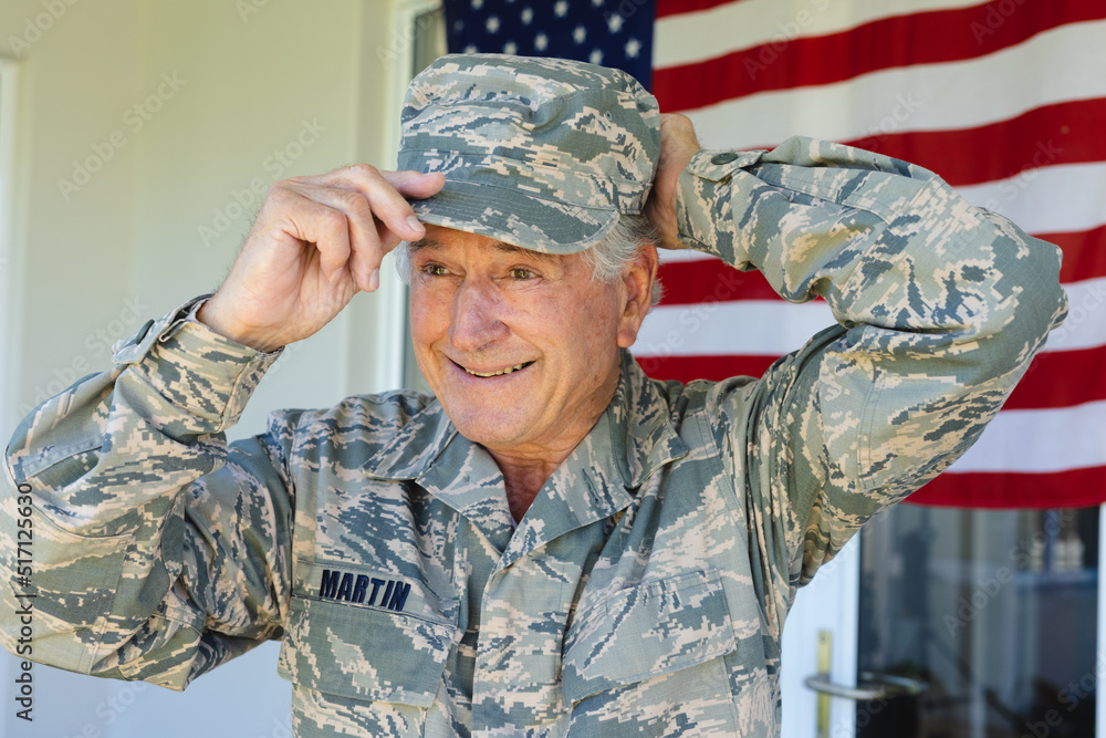 Fototapeta premium Smiling caucasian senior army soldier in camouflage clothing wearing cap against flag of america