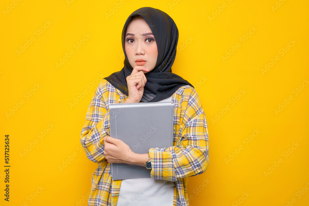 Pensive Young Asian Muslim student holding book, looks seriously thinking about a question isolated on yellow background. Back to school in high school college concept Pensive Young Asian Muslim student holding book, looks seriously thinking about a question isolated on yellow background. Back to school in high school college concept