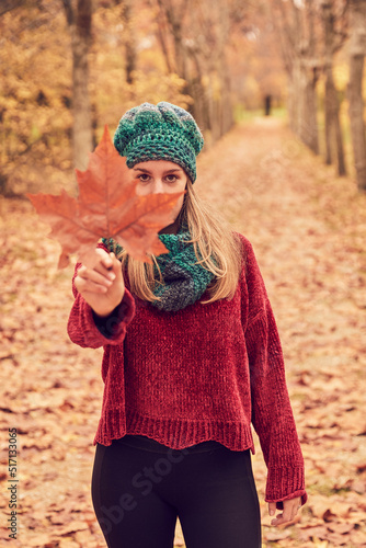 White Caucasian woman in blue cap and scarf and maroon sweater looking into camera clutching a brown five-pointed leaf covering her mouth and nose in a park in autumn