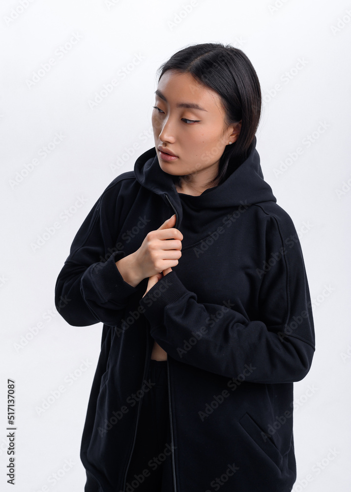 A beautiful Asian girl in a black casual tracksuit poses against a white wall in a photo studio A beautiful Asian girl in a black casual tracksuit poses against a white wall in a photo studio