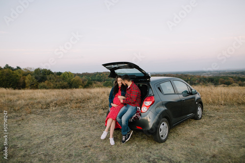 Young family sits in the trunk of a new car. Future parents laugh and hug dressed in casual clothes of red colors. Pregnant expectant mother in a dress in autumn meadow. High quality shot.