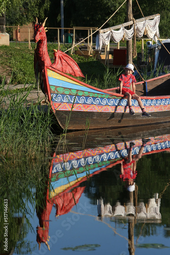 Happy tourist child, kid, boy in Suzdal town, Vladimir region, Russia. Suzdal architecture, landmark, monument. Suzdal sightseeing. Boat with dragon and river. Russian style. Reflection on water, view