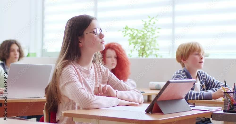 Cute female student in eyeglasses with long brown hair raising hand and waiting for turn to answer tricky questions. Children sitting at desks with laptops and tablets in classroom. Cute female student in eyeglasses with long brown hair raising hand and waiting for turn to answer tricky questions. Children sitting at desks with laptops and tablets in classroom.