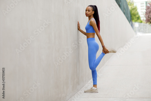 Side view of a fit black young woman stretching her legs in sports blue clothes against a gray wall.