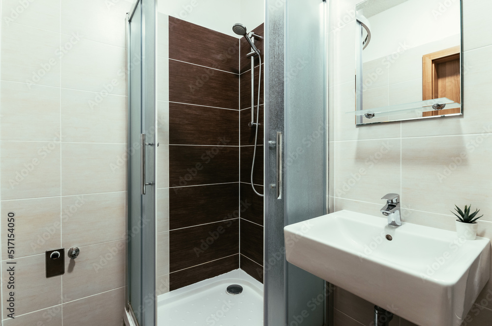 Obraz premium A small modern bathroom with a large shower, a square rectangular sink, a mirror with a glass shelf and on wall connection for a washing machine with water tap and water waste.