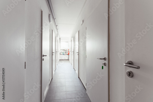 Hallway in a house with many solid white doors that have round handles of brushed metal. One door has a key in the lock. At the end of the corridor there are pipes and a window.