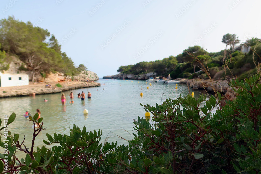 Cala en Blanes, Menorca (Minorca), Spain. Picturesque beach - Platja Cala en Blanes. View through the greenery, out of focus, unidentified people