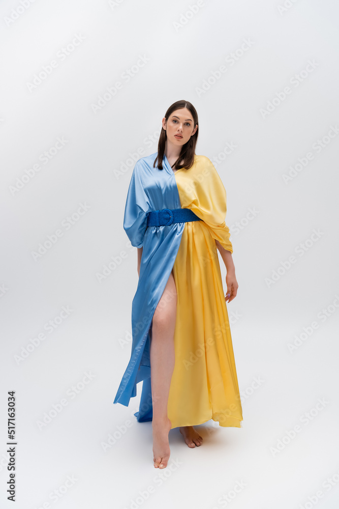 full length of barefoot young ukrainian woman in blue and yellow dress posing on grey.