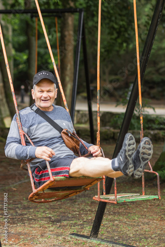 Carefree senior man in casual clothes and black cap having fun and swinging on swing in amusement park.