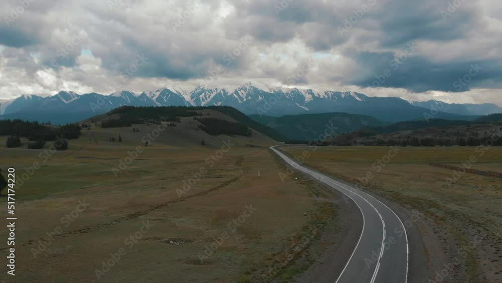 Aerial low air flight over mountain perfect asphalt highway road and meadow at sunny summer morning. Near green trees. Fast horizontal movement with sun rays. Snow mountains on background Aerial low air flight over mountain perfect asphalt highway road and meadow at sunny summer morning. Near green trees. Fast horizontal movement with sun rays. Snow mountains on background