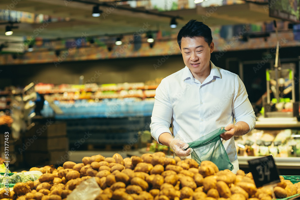 young asian man chooses and picks in eco bag vegetables potato or fruits in the supermarket. male customer standing a grocery store near the counter buys and throws in a reusable package in market