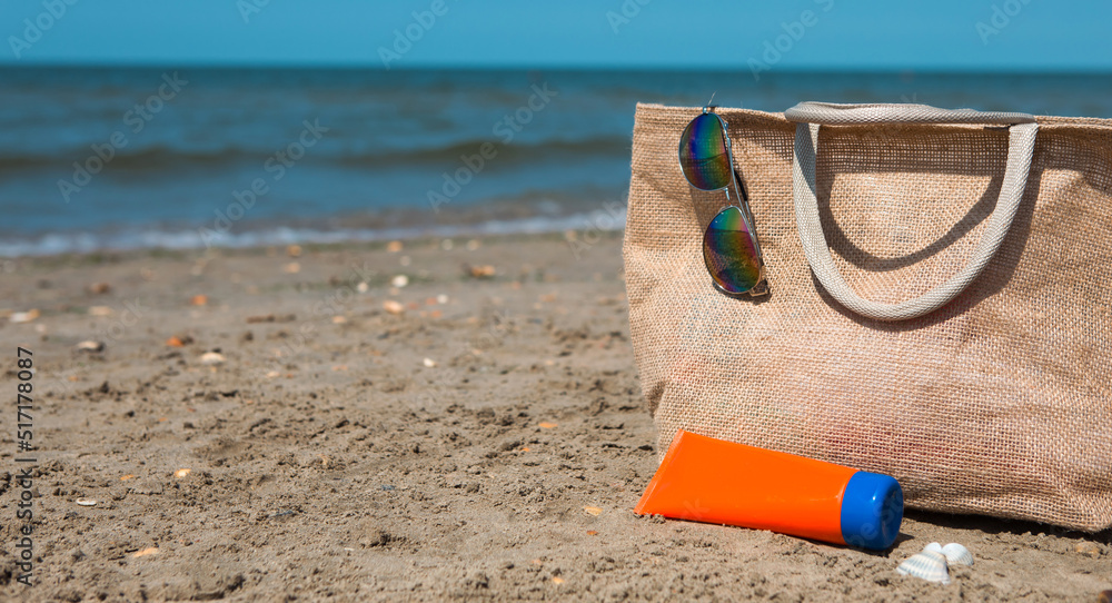 An orange tube with mock up sunscreen, sunglasses and a beach bag stand on the sand near the sea and swing from the breeze. UV protection, beach holidays, travel An orange tube with mock up sunscreen, sunglasses and a beach bag stand on the sand near the sea and swing from the breeze. UV protection, beach holidays, travel