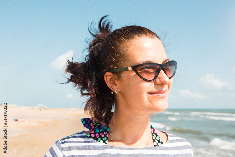 Portrait close-up happy brunette woman in a striped dress wearing sunglasses stands by the sea, looks at the waves, laughs and smiles. Tourism, beach holidays, breeze, journeys, travel