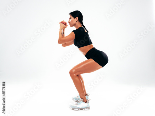 Fitness confident woman in black sports clothing. Sexy young beautiful model with perfect body. Female isolated in studio. Stretching out before training.Making squats on grey background. Full length