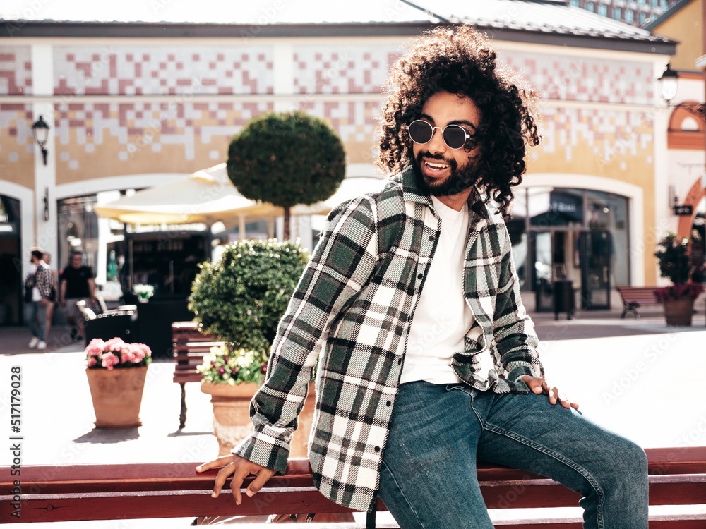 Handsome smiling hipster  unshaven Arabian man dressed in summer  clothes. Fashion male with long curly hairstyle posing in the street at   and happy. Sitting at the bench Stock Photo |