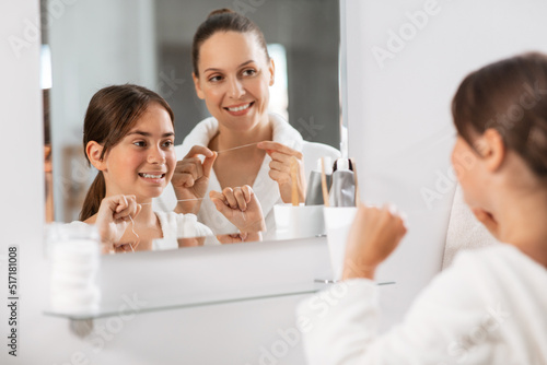 beauty, hygiene, morning and people concept - happy smiling mother and daughter with dental floss cleaning teeth and looking to mirror at bathroom