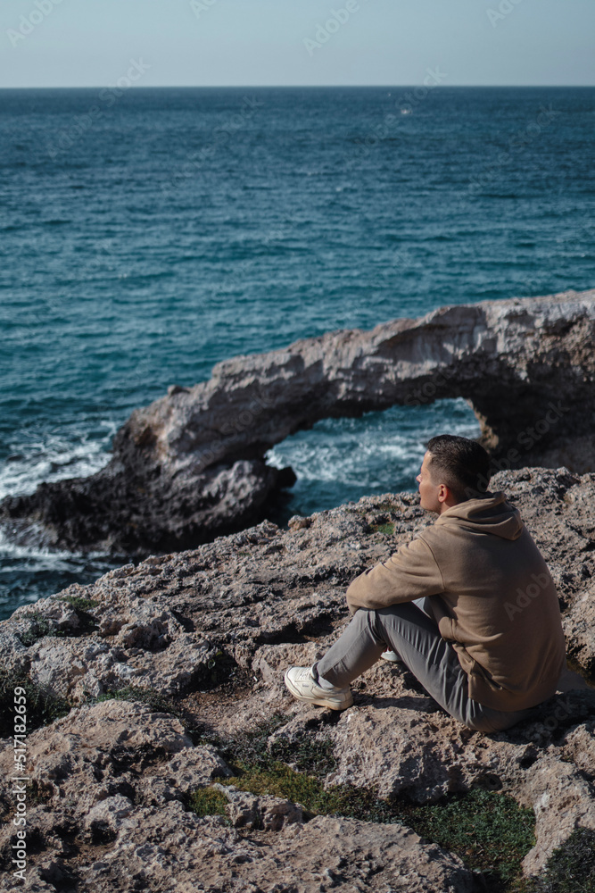 Man from behind is sitting at the edge and looking at sea cave arch bridge