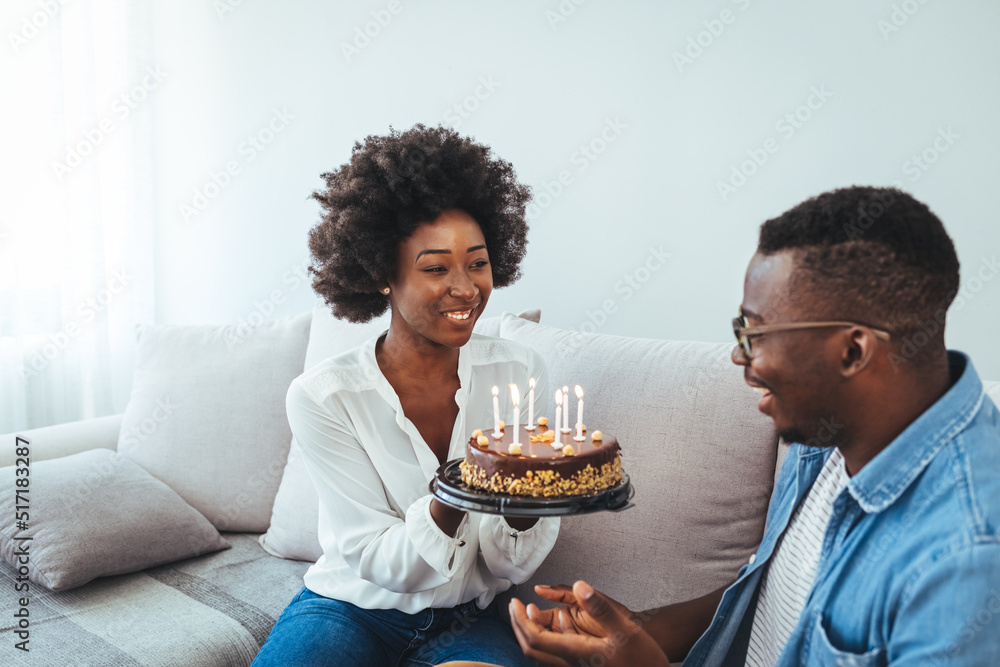 Multiethnic couple sitting at table and celebrating birthday at home in living room. Young african girl surprised on seeing birthday cake with her boyfriend. 