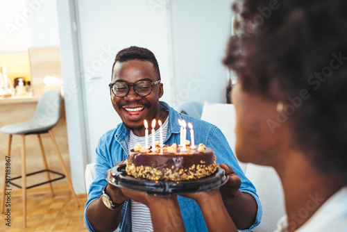 Happy African American couple celebrating anniversary or wife birthday at home. Portrait of happy woman blowing out candles on birthday cake standing with husband together in Living room