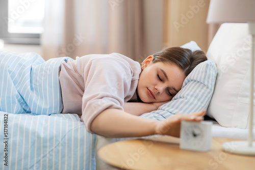 people, bedtime and rest concept - teenage girl sleeping in bed at home in morning and reaching to alarm clock