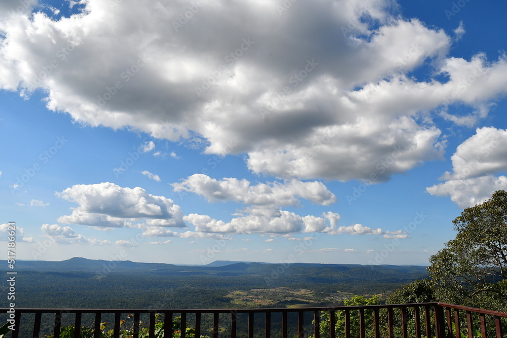 A photograph of a summer sky and clouds during the day. The atmosphere of the sky taken from a viewpoint with a long wooden railing and trees on the sides.

