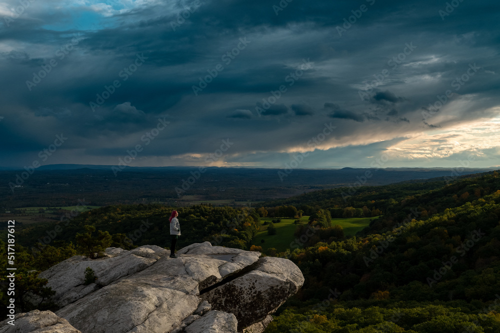 girl stand on edge of rock, aerial view of mountains, sunset light and cloudy sky, Gertrude's Nose NY