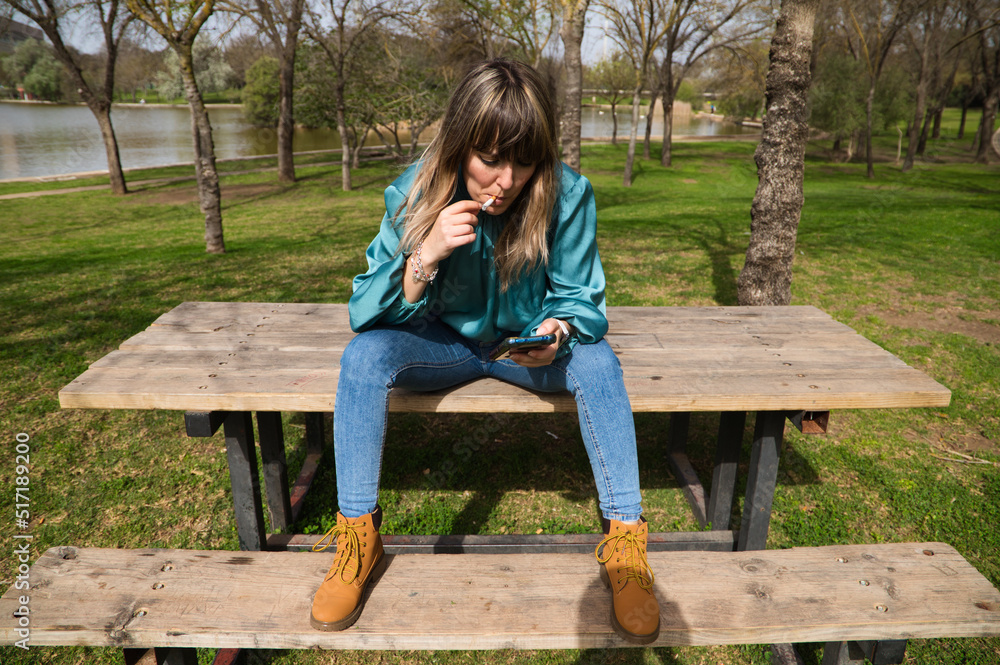 A young, pretty, blonde woman is sitting at a wooden table in the park, looking at her mobile phone and smoking a cigarette. In the background you can see the river on a sunny day. Stop smoking A young, pretty, blonde woman is sitting at a wooden table in the park, looking at her mobile phone and smoking a cigarette. In the background you can see the river on a sunny day. Stop smoking