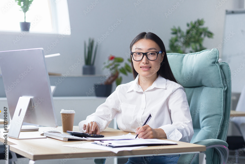 Workaholic. A young Asian business woman, sitting at the table drinking coffee to go, a paper cup of coffee is standing on the desktop near the computer. He looks at the camera, smiles. Workaholic. A young Asian business woman, sitting at the table drinking coffee to go, a paper cup of coffee is standing on the desktop near the computer. He looks at the camera, smiles.