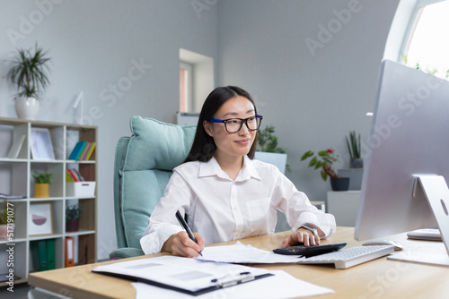 Work with documents. Portrait of a young beautiful business woman Asian accountant works with documents and reports. Sitting at the desk in the office, writing. © Liubomir