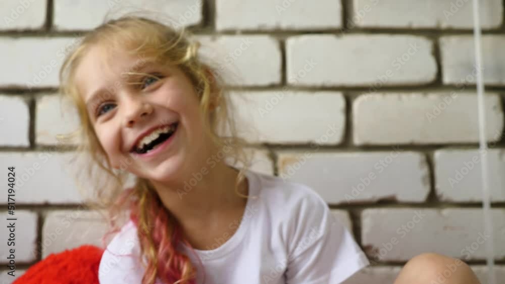 A beautiful blonde girl with dyed red hair tips, laughing against a white brick wall. The child was watching a video on her smartphone. Slow motion. Selective focus A beautiful blonde girl with dyed red hair tips, laughing against a white brick wall. The child was watching a video on her smartphone. Slow motion. Selective focus