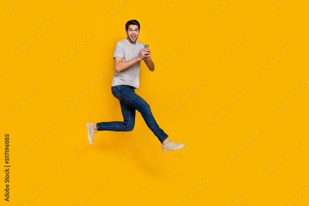 Full length photo of nice brunet millennial guy jump with telephone wear t-shirt jeans sneakers isolated on yellow background