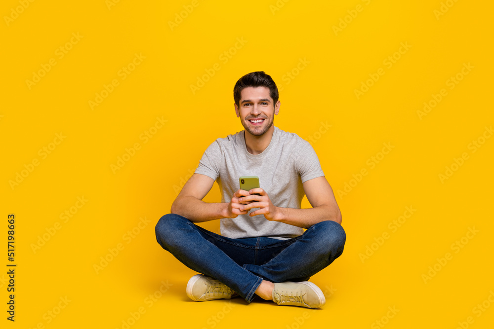 Full size photo of funny brunet young guy sit hold telephone wear t-shirt jeans sneakers isolated on yellow background Full size photo of funny brunet young guy sit hold telephone wear t-shirt jeans sneakers isolated on yellow background