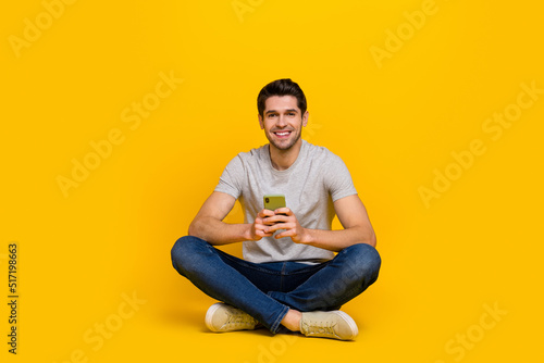 Full size photo of funny brunet young guy sit hold telephone wear t-shirt jeans sneakers isolated on yellow background © deagreez
