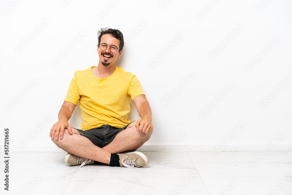 Fototapeta premium Young caucasian man sitting on the floor isolated on white background with glasses and happy