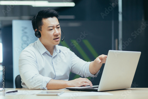 A young handsome Asian businessman in headphones with a microphone sits at a laptop in the office, conducts an online conference, a remote meeting with clients, partners. Tells, explains, smiles