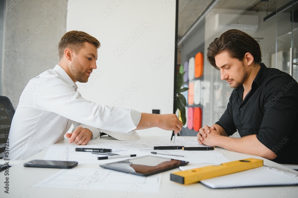 Two young architects with blueprints of a house standing in office, talking. Two young architects with blueprints of a house standing in office, talking.