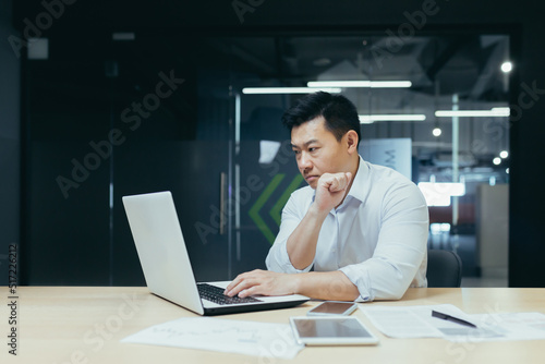 A young handsome man, an Asian businessman, is working with a laptop, typing. Sitting at a desk in a modern office