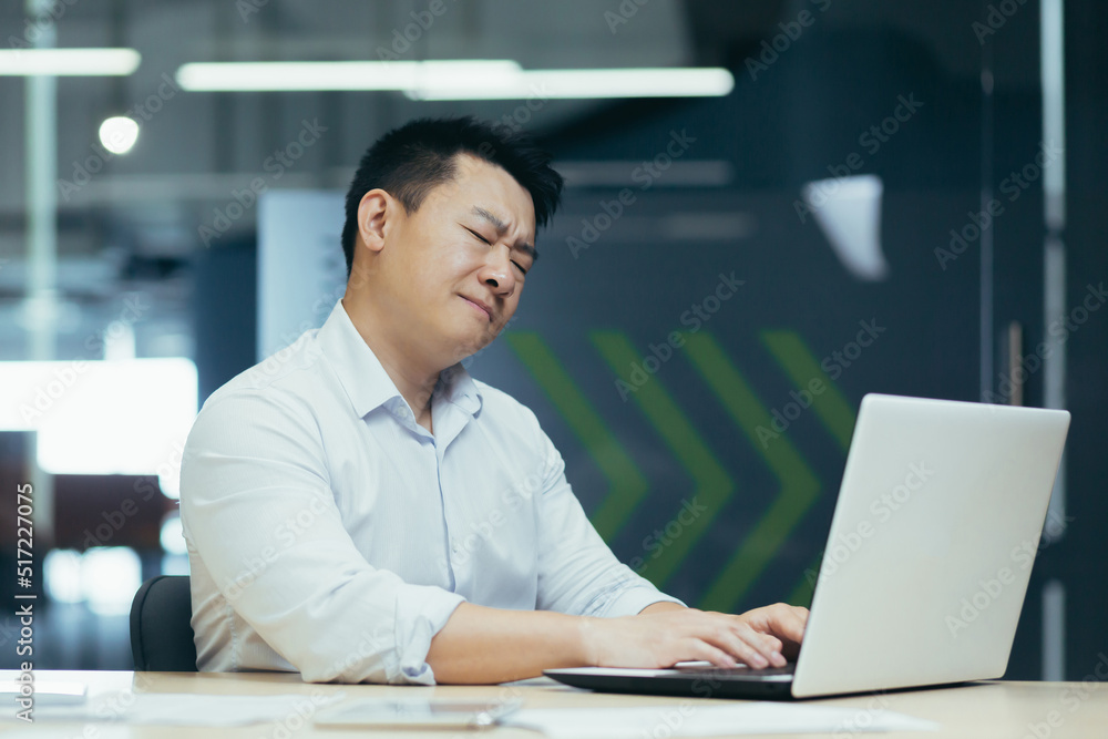 A young man, an Asian businessman at work in the office, sits at the table, holds his neck, feels pain, closes his eyes, A young man, an Asian businessman at work in the office, sits at the table, holds his neck, feels pain, closes his eyes,
