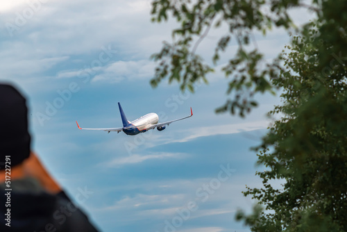 rear view on a passenger airliner taking off in the blue sky against the background of foliage of trees and the silhouette of a girl looking into the distance