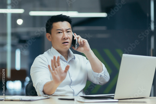 Worried and serious young male Asian businessman talking on the phone in the office. Sitting at a table with a laptop and documents. Trying to solve problems © Liubomir
