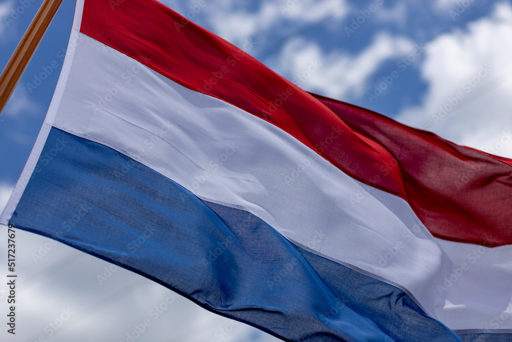 Naklejka premium Brightly lit tricolor of Dutch national flag blowing in a strong wind with texture and detail in the folds against a blue sky with clouds