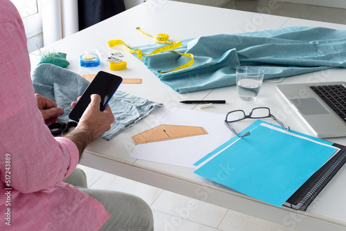 clothing designer sitting in his studio, sewing workshop. holding mobile, phone, smartphone. horizontal