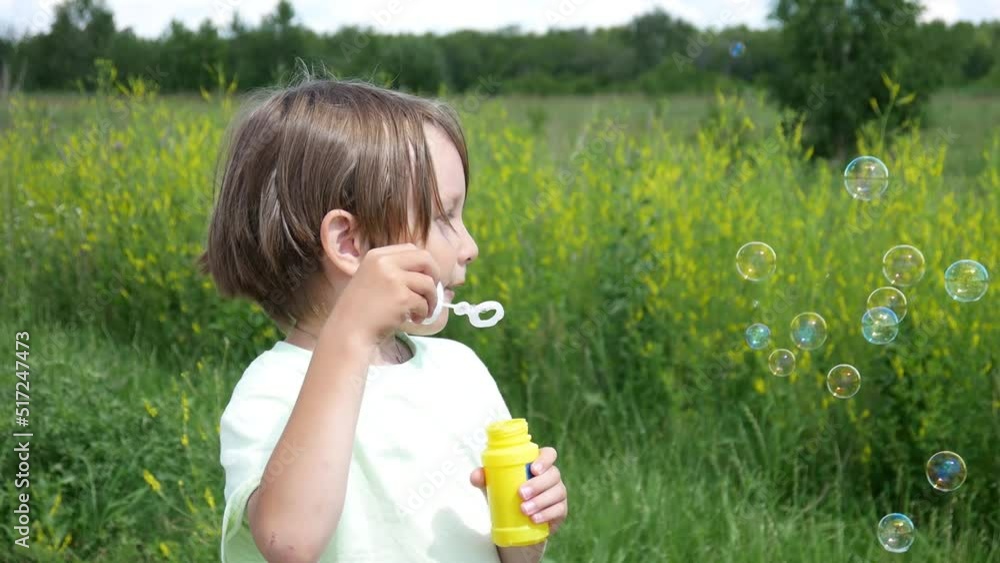 A boy blowing soap bubbles standing on the side of a green field