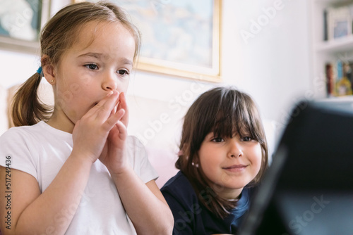 Two little girls attentively watching a video on a tablet in their living room with astonished faces. Concept of childhood, technology, learning, having fun, the Internet and connectivity.