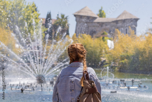 A woman with pigtails looks at the fountain gushing from the lake against the backdrop of trees and unusual park architecture. Selective focus.